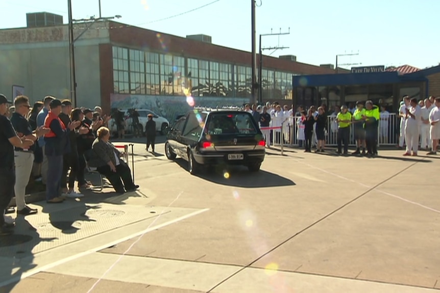 Workers outside a factory with a hearse in the middle