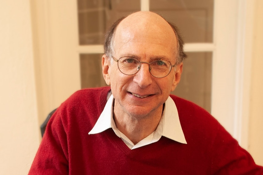 A man, wearing glasses, a red jumper, and white-collared shirt, smiles.