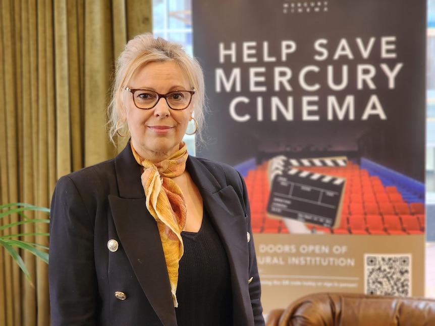 A woman with blonde hair standing next to a sign saying HELP SAVE MERCURY CINEMA