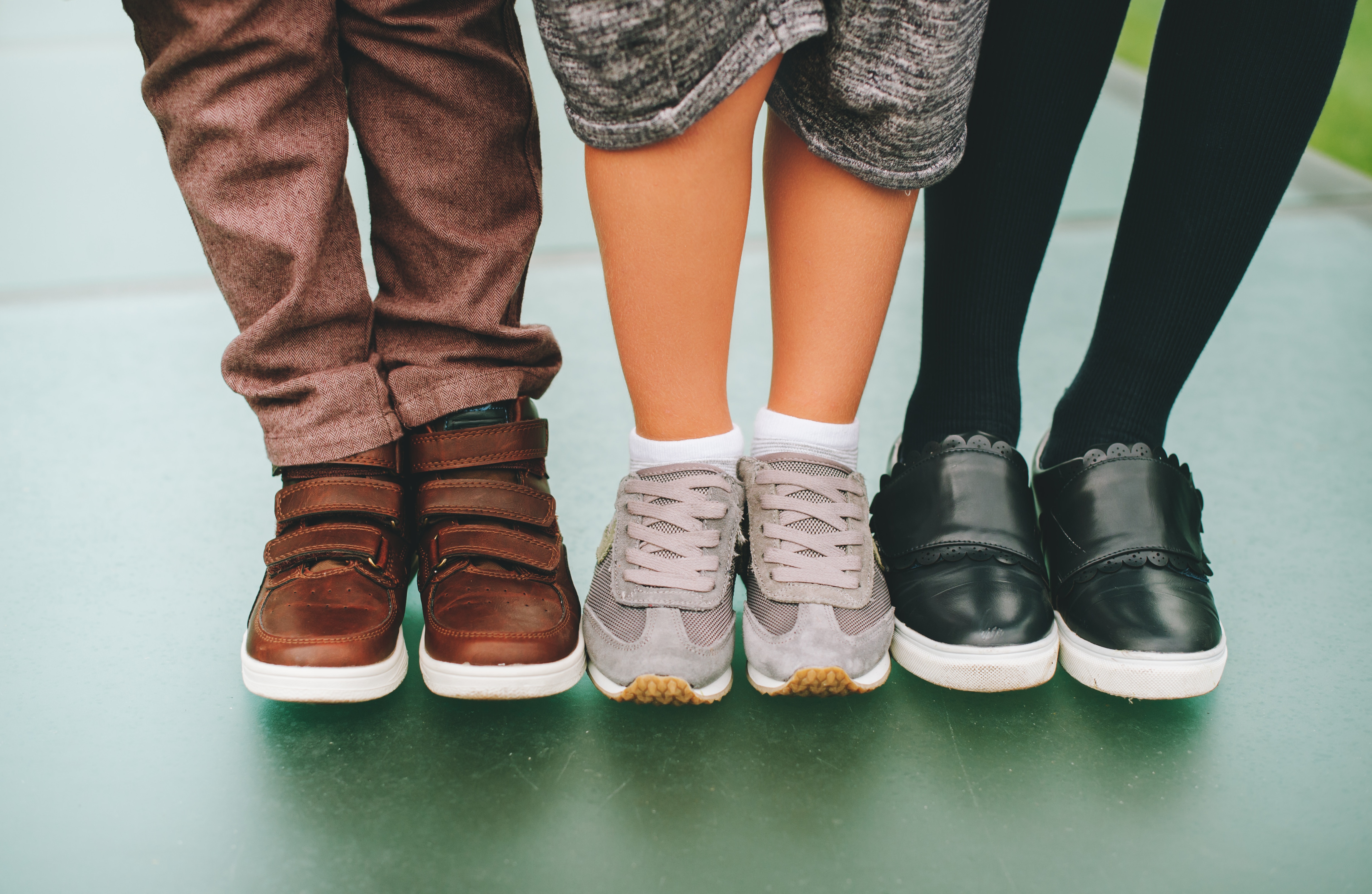 Three pairs of children's feet wearing, respectively, brown shoes with velcro straps, grey cross trainers and black leather. 