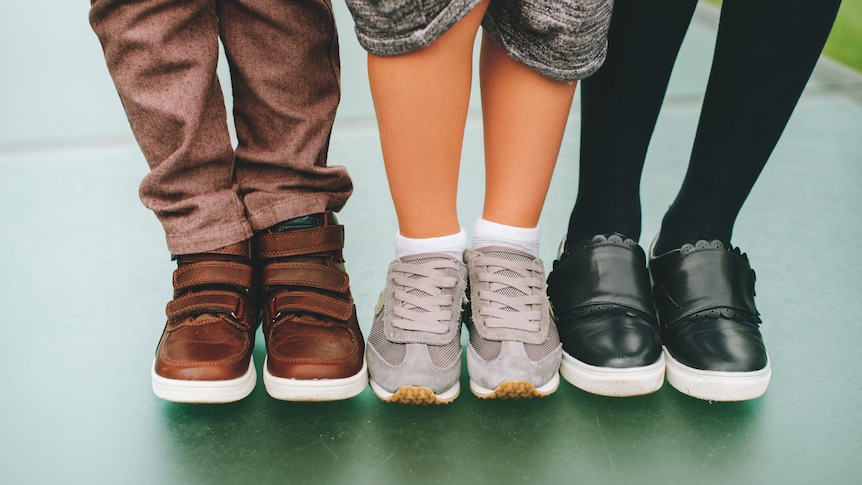 Three pairs of children's feet wearing, respectively, brown shoes with velcro straps, grey cross trainers and black leather. 