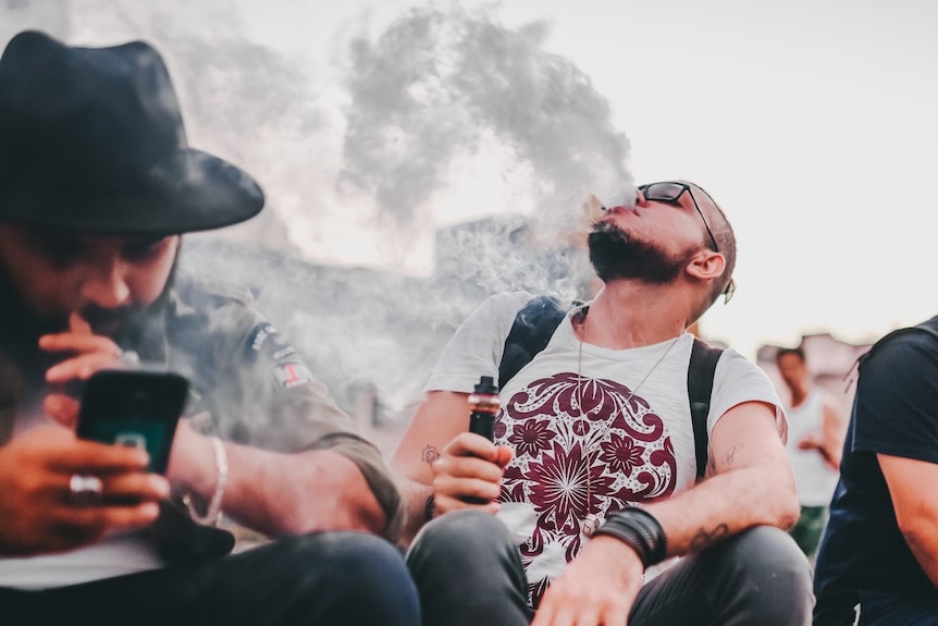 Man exhaling e-cigarette vapour into the air, right, into the face of another man, left