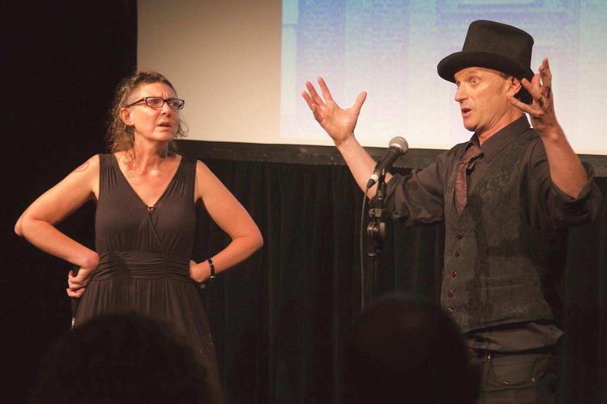 A man, with a top hat and hands thrown into the air, watched by a woman, hands on hips, stand on stage near microphones.