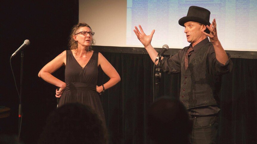 A man, with a top hat and hands thrown into the air, watched by a woman, hands on hips, stand on stage near microphones.
