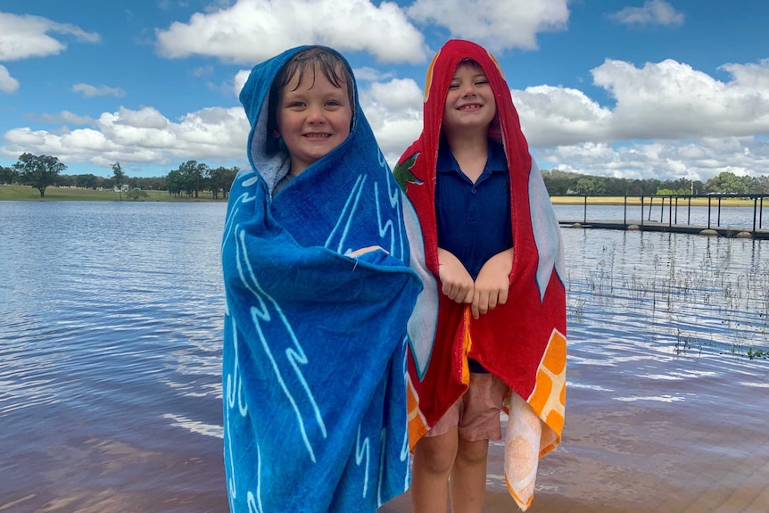 Two boys standing near a full dam with hooded towels on.