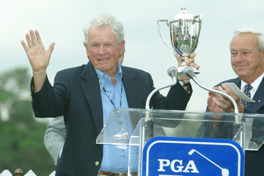 A medium shot of an older man waving with one hand and holding up a trophy with the other during an outdoor PGA prize ceremony.