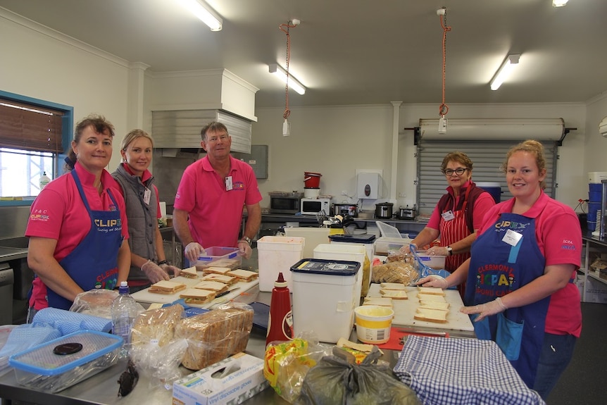 A wide shot in the kitchen with sandwiches being prepared by  five volunteers in pink shorts and blue aprons.