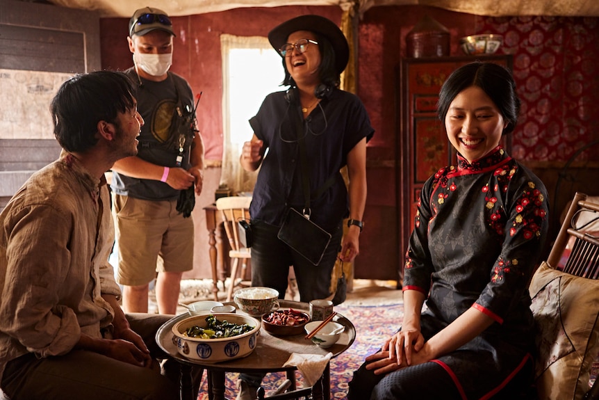 In an 19th century room, a young Asian woman in dark clothes laughs with two young Asian actors sitting down. Masked man in bg