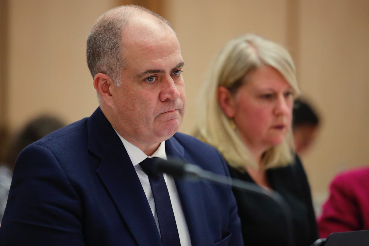 David Anderson reacting to questions at Senate Estimates. He is a balding white man with blue eyes, wearing a blue suit. 