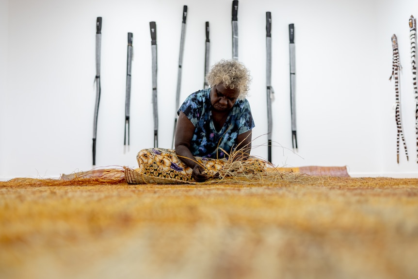 Doreen Jinggarrabarra, a Burarra woman with dark brown skin and grey curly hair sits in a gallery weaving a large mat.