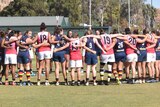 Women in AFL uniforms standing in a circle huddle with their arms around each other on a football oval.