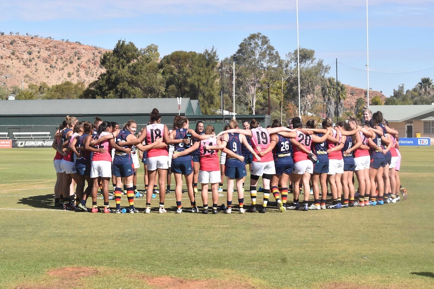 Women in AFL uniforms standing in a circle huddle with their arms around each other on a football oval.