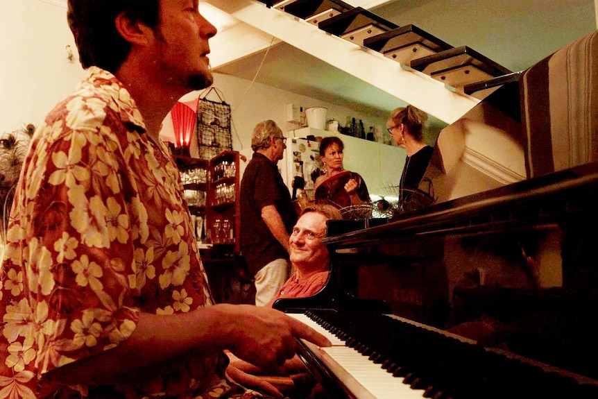 Man in floral shirt in living room playing piano at a party. Man smiling watching. People talking