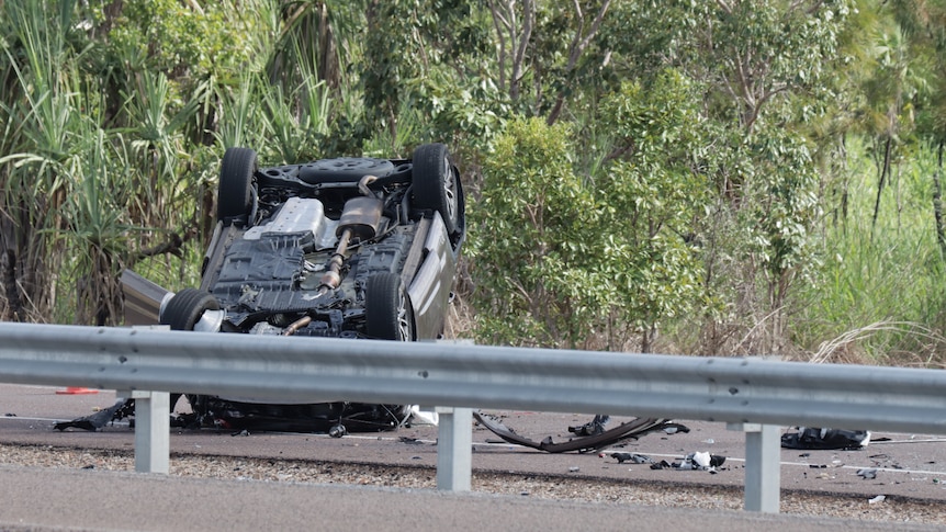 A crashed car rested on its roof on the side of a highway, in front of scrub. 