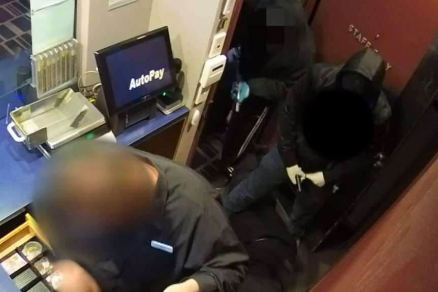A staff member handing over cash to two armed men, dressed in all black during an aggravated robbery.