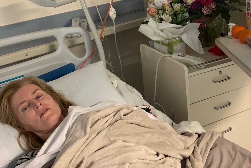 Woman lying in hospital bed looking very sick with flowers on side table.