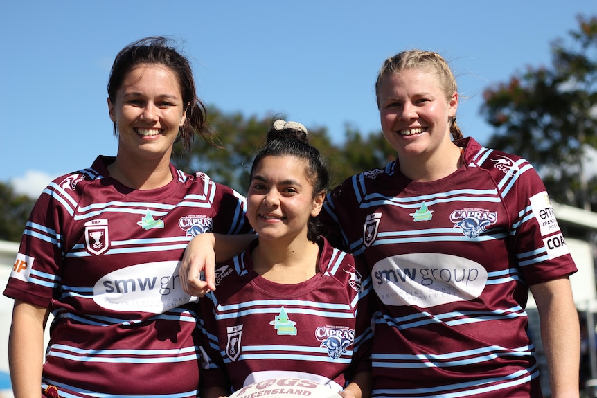 Three women in maroon and blue jerseys smiling on a rugby pitch.