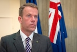 A man in a grey suit stands in front of an Australian flag.