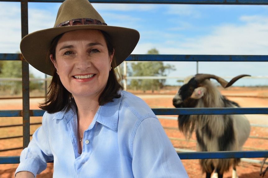 Prue Adams wearing wide brimmed hat standing in front of goat in a yard.