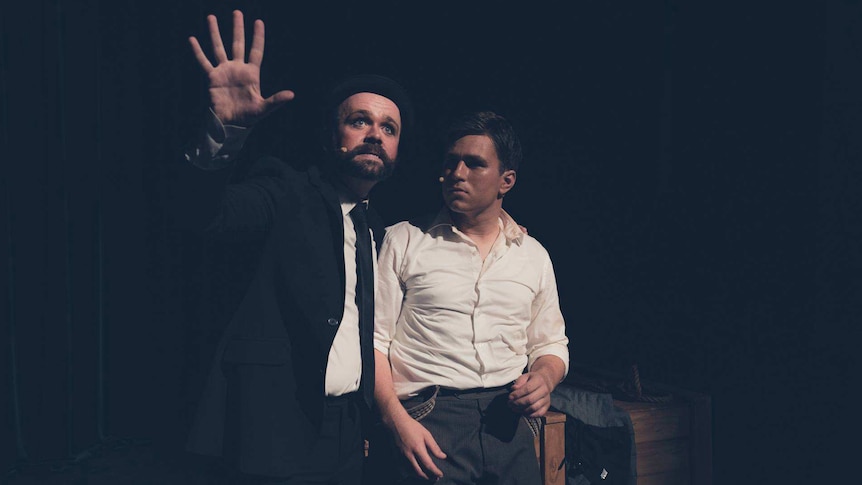 Two male performers stand on dark stage, one in black suit the other in white shirt in dramatic character pose.