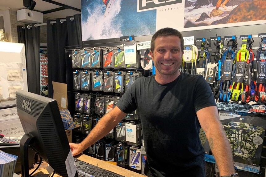 A man in a black shirt smiles behind the counter of a surf shop