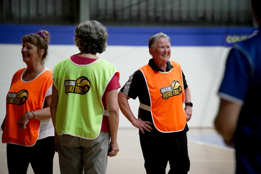 Three women wear basketball bibs at a basketball court in Canberra.  They are smiling and playing walking basketball. 