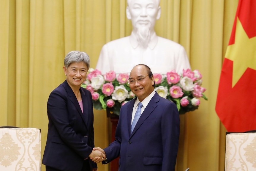Penny Wong and Nguyen Xuan Phuc shake hands in front of a white bust of Ho Chi Minh with Vietnamese flag of yellow star on red 