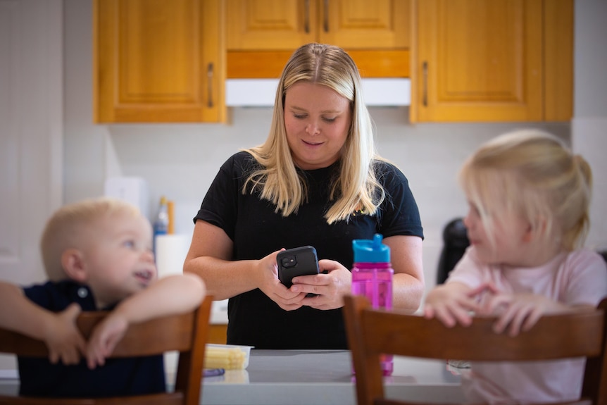 Amy looking seriously at her phone in the kitchen, while two young children lean on wooden chairs smiling the foreground.