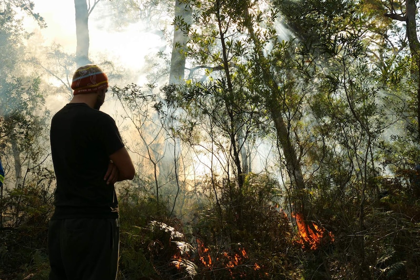 Young man in beanie standing with arms folded watching a small area of bracken burning
