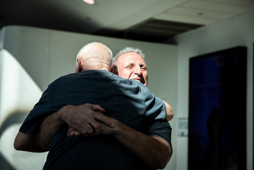 Two older men hug one another, with one's back to the camera. 