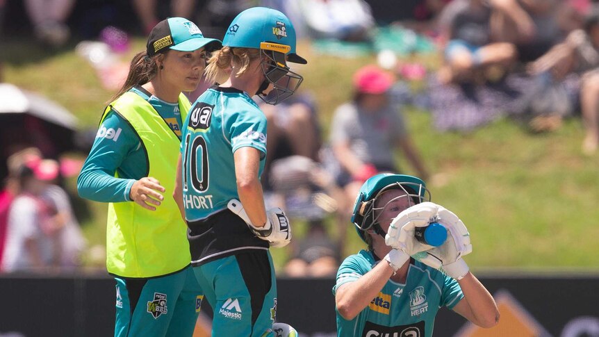 Two cricketers, one with a high-vis vest over her uniform, stand next to another kneeling and drinking from a water bottle.