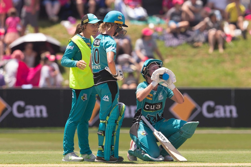 Two cricketers, one with a high-vis vest over her uniform, stand next to another kneeling and drinking from a water bottle.