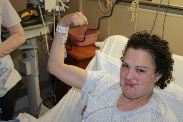 A woman in a hospital bed holds up her arm in which a drip is inserted.