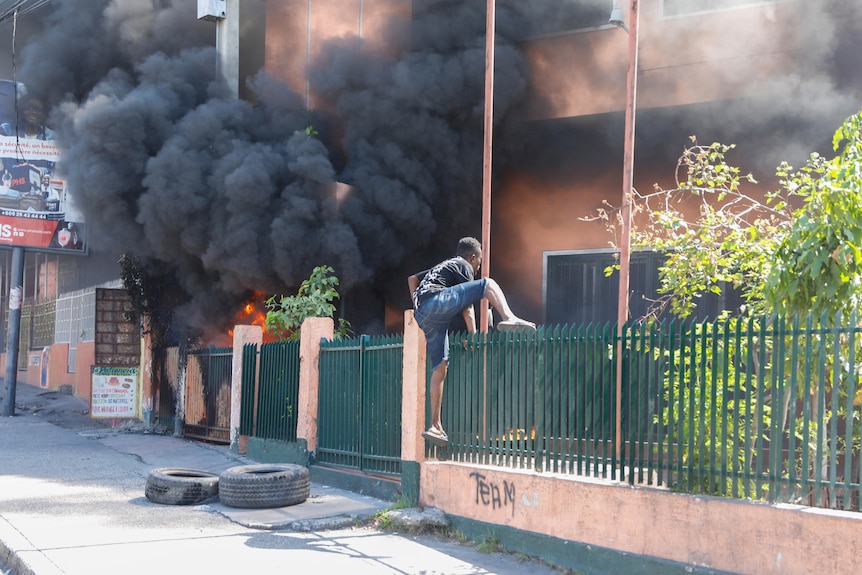 A man climbs over the fence of an office building that has large plumes of black smoke billowing out the windows.