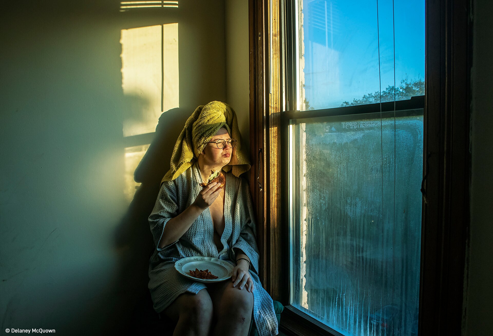 A woman in a bathrobe and towel wrapped around her hair sits at a window and eats a sandwich. 