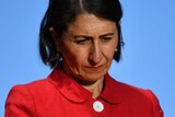 Gladys Berejiklian wears a red jacket and looks down towards the floor at a press conference