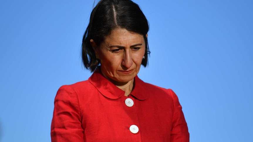 Gladys Berejiklian wears a red jacket and looks down towards the floor at a press conference