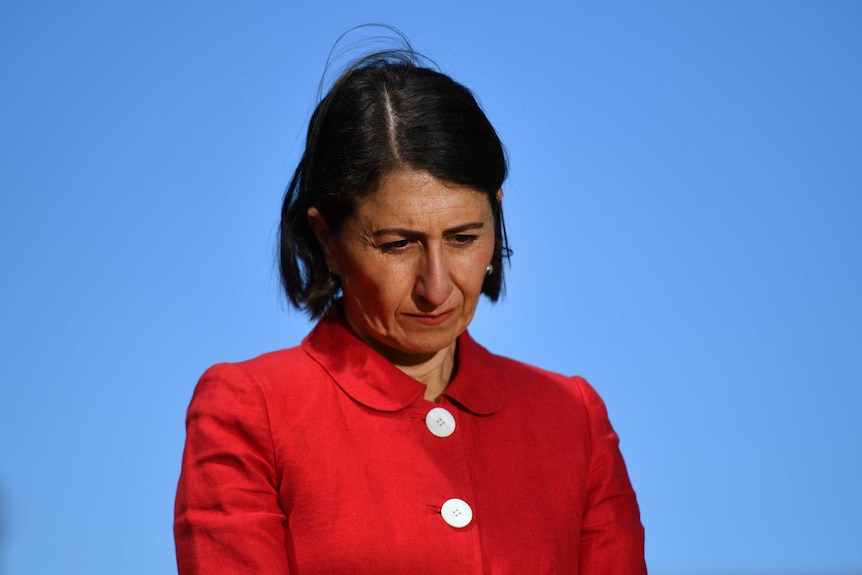 Gladys Berejiklian wears a red jacket and looks down towards the floor at a press conference