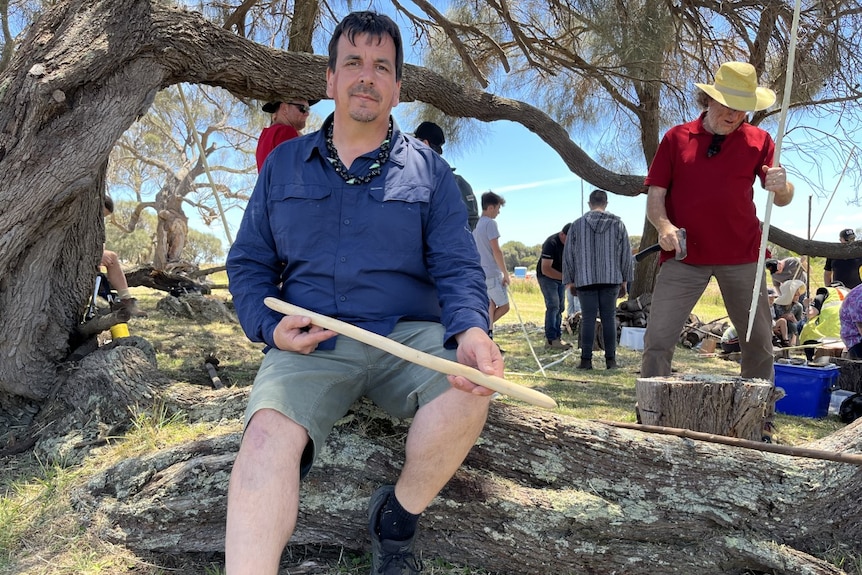 A man, holds a stick while he sits on a log