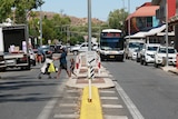 A street in the Alice Springs CBD. A family crosses the road, pushing a stroller. A bus is coming in the other lane.