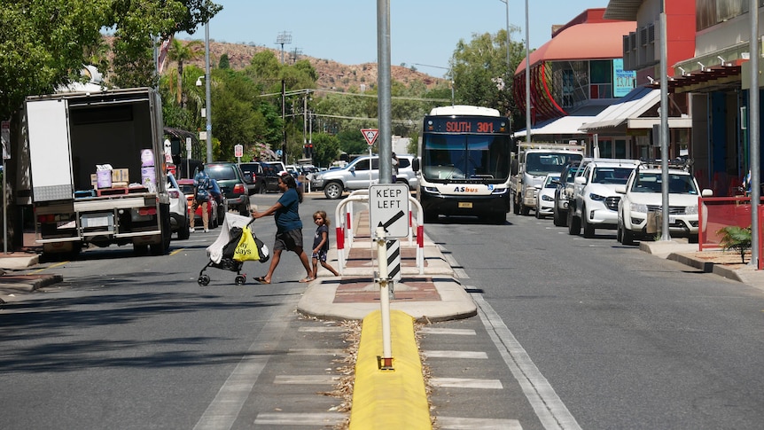 A street in the Alice Springs CBD. A family crosses the road, pushing a stroller. A bus is coming in the other lane.