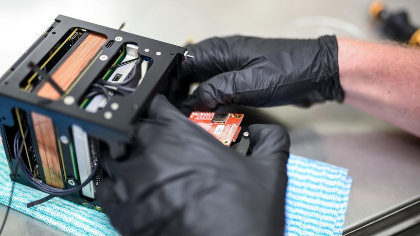 A person wearing black plastic gloves works on a cube shaped spacecraft