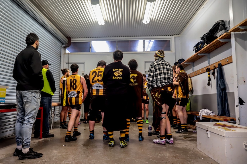 Male football players wearing gold and brown jerseys and robes in fluorescent lit tin shed changerooms