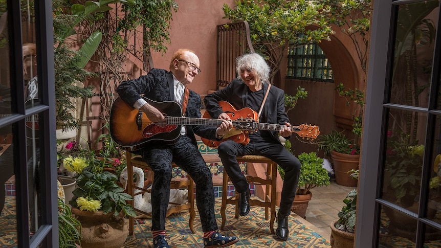 Peter Asher and Albert Lee sit in garden playing guitar, wearing black suits, surrounded by green plants and trees.