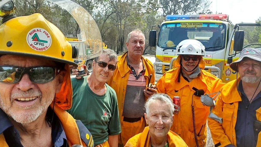 John Foster takes selfie photo of Woodgate firefighters all aged over 70, in uniform with fire trucks in the background.