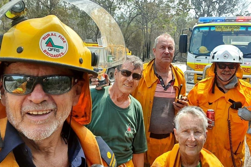John Foster (left) takes selfie photo of Woodgate firefighters all aged over 70, in uniform with fire trucks in the background.