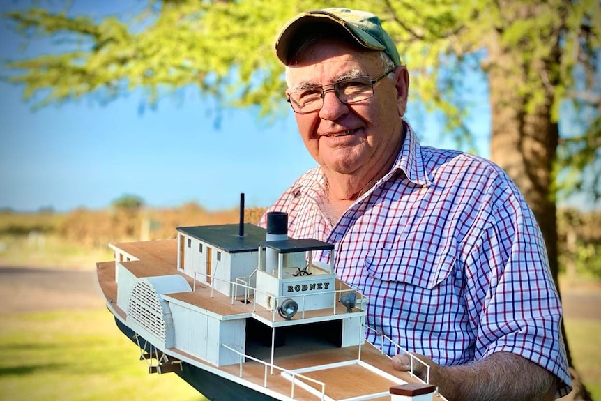 Man in checked shirt and cap holding a scale model of the PS Rodney