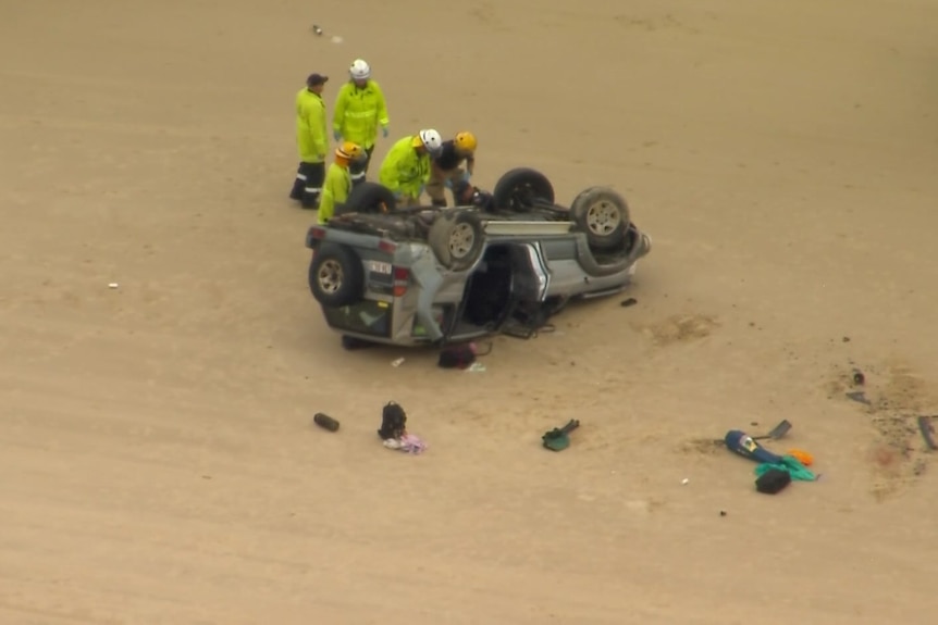 An aerial view of an upside-down four-wheel-drive vehicle on a beach, with emergency services workers clustered around it. 