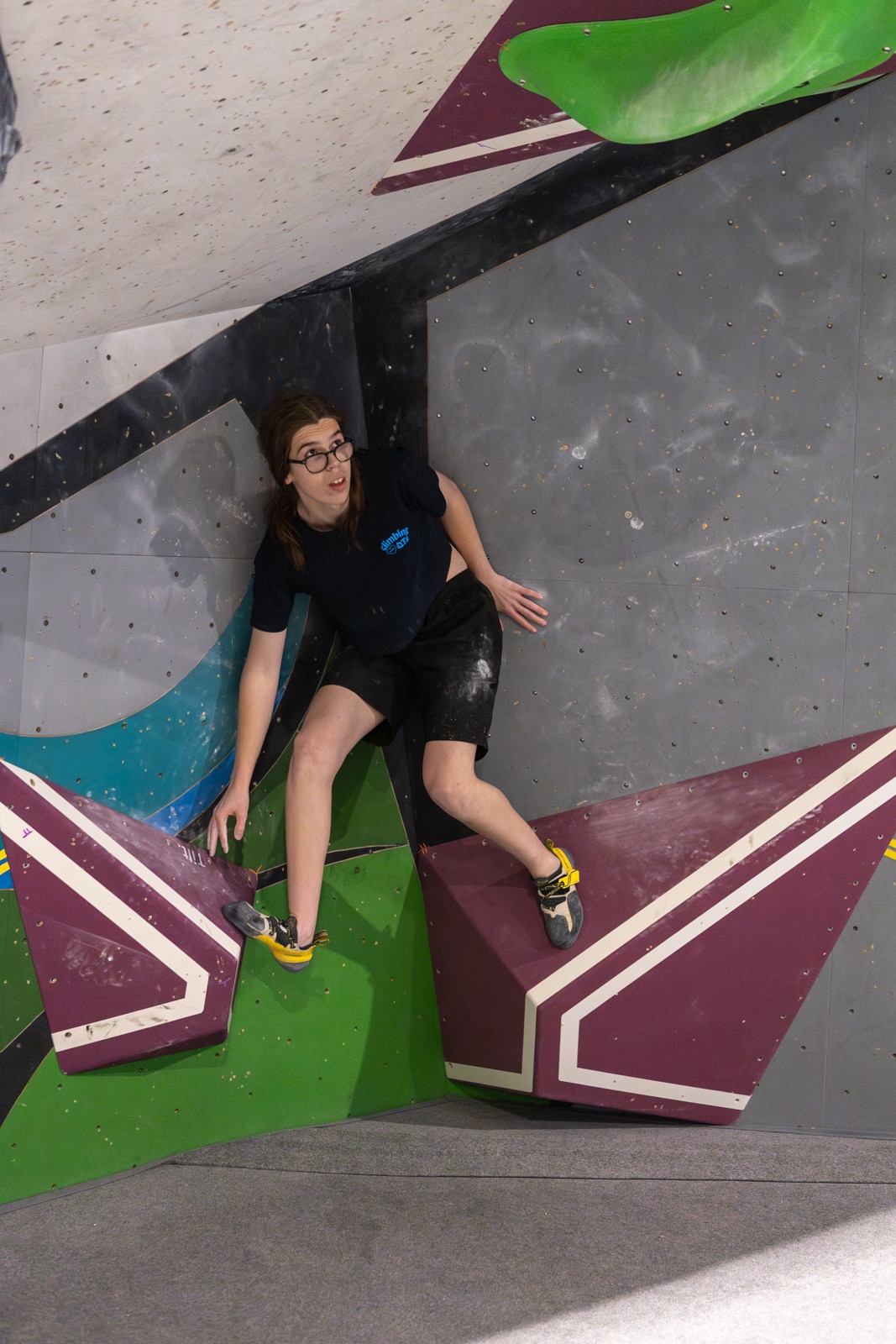 A dark-haired young woman in glasses on an indoor rock-climbing wall.