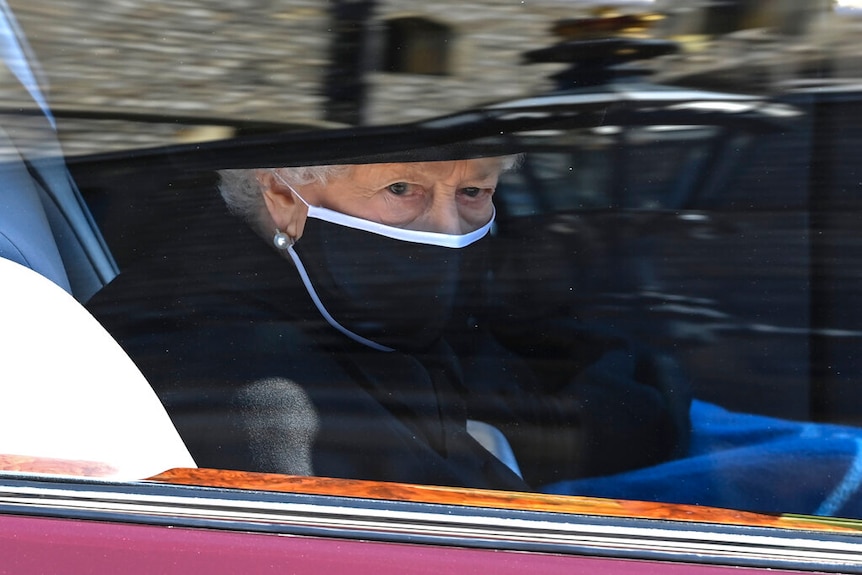 You view a closeup of the Queen wearing a black face mask and hat through the window of a car.